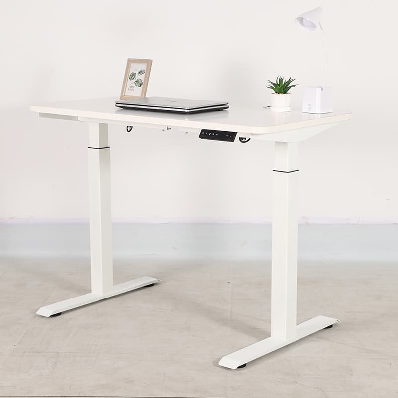 Escritorio de pie ajustable 2B2 blanco con computadora port&aacute;til, planta y l&aacute;mpara sobre un piso gris claro contra una pared blanca.